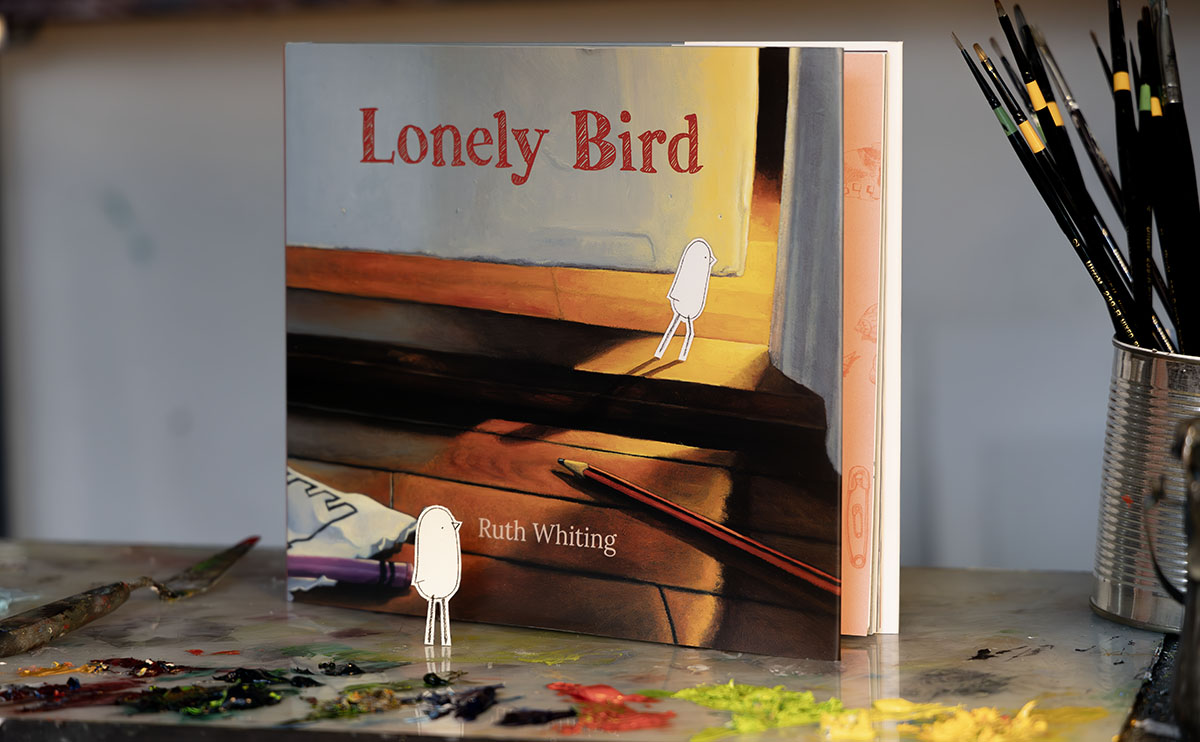 image of Lonely Bird, a children's book painted by Ruth Whiting, shown here on top of a pallet of oil paint with the character Lonely Bird looking at the book